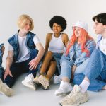 Group of cheerful diverse teenager friends talking to each other while sitting together on floor against white background — A