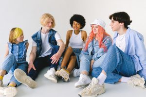 Group of cheerful diverse teenager friends talking to each other while sitting together on floor against white background — A