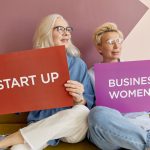 Two women on a sofa holding signs promoting startup and business women. — Cape Town Florist - Luxury Floral Designs Crafted b